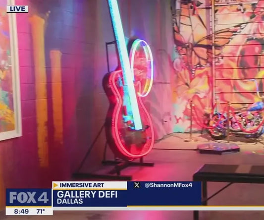 A neon colored guitar with led lights of pink and blue. A news heading underneath showcases the Gallery Defi on Fox News broadcast, with images of butterflies in the back of the room it is spaced in for the 3d guitar art.