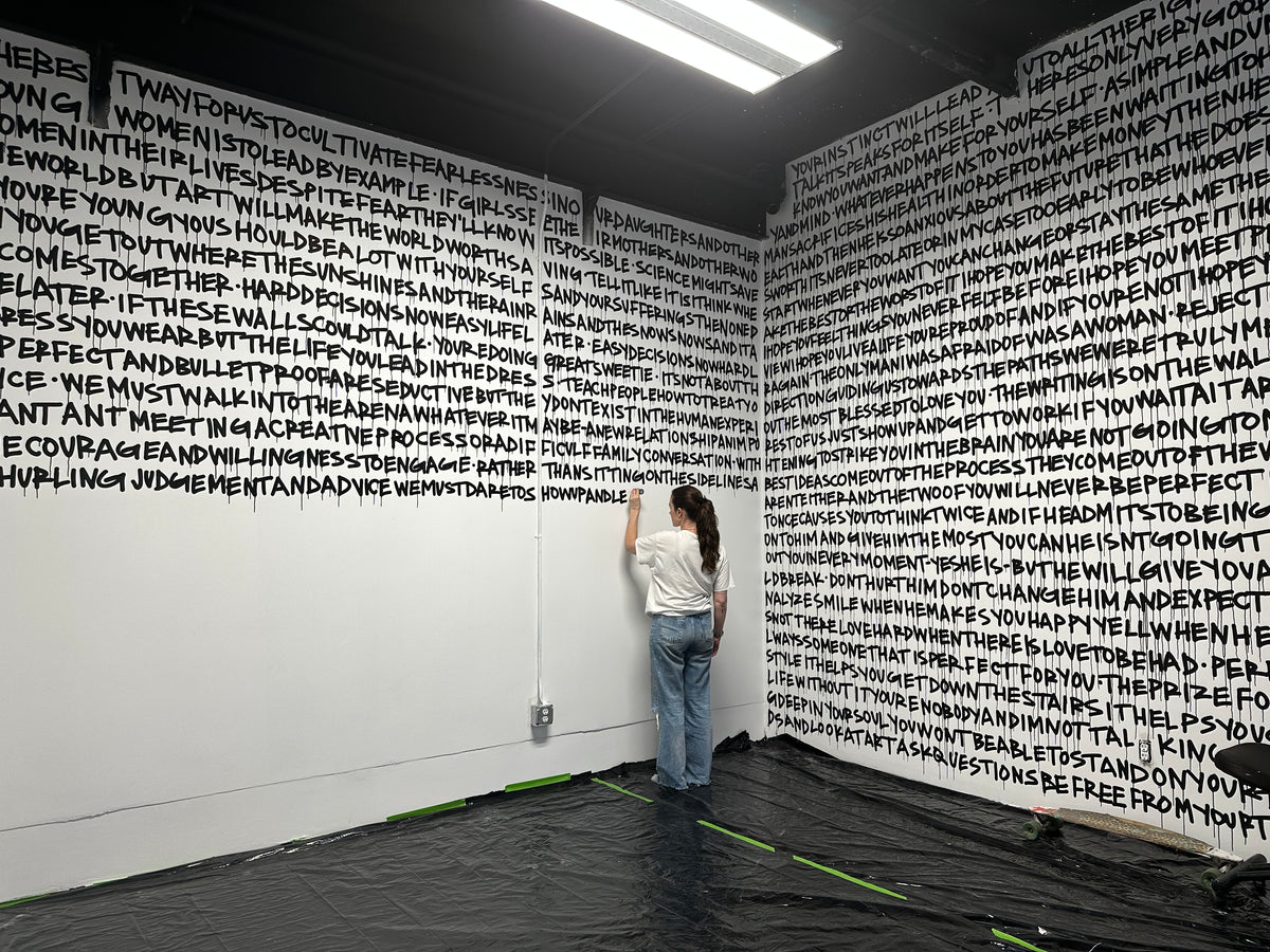 Leah Kirsch painting black handwritten text across the white walls of her room at Gallery DeFi
